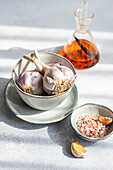 From above of raw garlic bulbs nestled in a bowl accompanied by a glass decanter filled with amber liquid and a smaller dish of pink salt all set on a sunlit surface