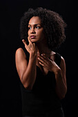 Hispanic young female with curly hair touching face and looking away while standing on black background