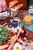 A hand pours coffee into a blue and white cup, part of a gourmet brunch spread including croissants, meats, cheese, and daisies.