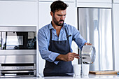 Focused bearded adult male chef in apron looking down while standing in kitchen and pouring ingredients from saucepan into glass during breakfast preparation in modern kitchen in daylight