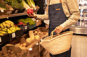 Cropped unrecognizable male seller in apron choosing fresh ripe bell pepper standing near assorted vegetables and fruits with straw basket in supermarket