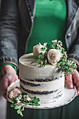 Unrecognizable female in green dress and leather jacket demonstrating appetizing cake decorated with flowers and sprigs on wedding day