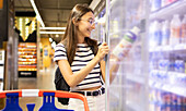 Smiling young female customer in casual clothes choosing bottle of fresh juice from fridge while shopping at supermarket