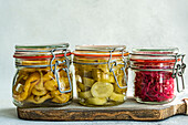 Three glass jars filled with fermented cabbage, spicy peppers, and cucumbers on a rustic wooden board against a gray background.