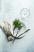 Draufsicht auf Tischdekoration mit frischer Pistazienpflanze auf Teller mit Besteck neben Serviette und Glas vor grauer Fläche im Tageslicht
