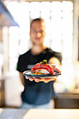 Ein nicht erkennbarer junger Mann mit gelbem Stirnband steht in der Küche und bietet mit ausgestreckter Hand appetitlich garnierte Sushi auf einem Teller an, während er in die Kamera schaut