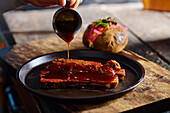 High angle of crop anonymous cook pouring delicious sauce on grilled pork ribs served on plate garnished with baked potatoes