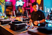 Chopstick with folded black napkin and empty drinking glasses arranged on neat wooden table at modern illuminated sushi bar against blurred male cook in black uniform