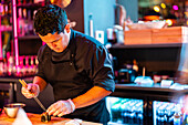 Focused male chef in black uniform and gloves using chopsticks for making fresh sushi roll while working in kitchen at illuminated modern nightclub