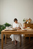 Full body of young female in gown sitting at wooden table with bowl of strawberries and glass bottles while making strawberry jam in room