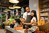 Happy young multiracial coworkers in aprons smiling and using monitor to enter information while standing at counter together during work