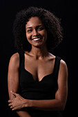 Smiling young Hispanic female model with Afro hairstyle in black clothes looking at camera while standing against black background