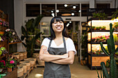 Happy young Asian florist in apron smiling and looking at the camera while standing with her arms crossed while working in a florist shop