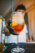 Unrecognizable bartender with gloves holding an orange cocktail with ice and a straw standing at bar in blurred background