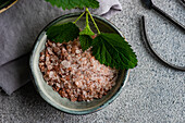 Close-up from above of vegan cooking setting featuring a bowl of Himalayan pink salt garnished with a fresh nettle leaf on textured gray backdrop with a pair of scissors nearby