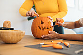 Anonymous women near table carving with tool scary teeth on orange pumpkin and preparing for Halloween decoration in day