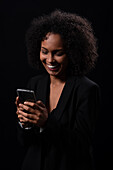 Positive Hispanic young female in outfit smiling and browsing mobile phone while standing on black background