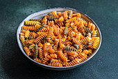 From above raw uncooked fusilli pasta made from wheat, beetroot and spinach in the bowl on dark green background