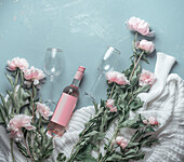 Flasche Rosenwein mit Weingläsern auf blauem Hintergrund mit weißem Strickpullover und vielen rosa Pfingstrosenblüten. Ansicht von oben. Schönheit