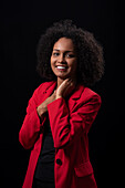 Smiling young Hispanic female model with Afro hairstyle in red coat looking at camera while standing against black background