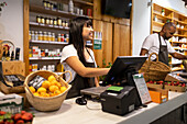 Cheerful Asian female seller in apron smiling and using computer while standing at counter with her colleague and various products while working in a supermarket