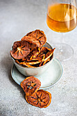 High angle of slices of dried apples served in ceramic bowl on plate near glass of juice against blurred gray background