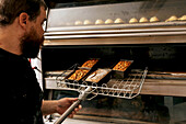 Männlicher konzentrierter Koch in Freizeitkleidung, der gebackenes knuspriges heißes Brot und Gebäck mit metallischem Schulterblatt aus dem Ofen in der Backstube nimmt
