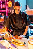 Confident focused young female cook in black uniform and gloves using blow torch while cooking fresh sushi in plate at in kitchen at restaurant
