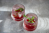 From above of crystal glasses with gin tonic and pomegranate garnished with rosemary, casting soft shadows on a grey concrete surface