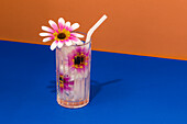 Transparent glass of refreshing cold drink decorated with pink flowers and straw placed on blue surface against bright orange wall