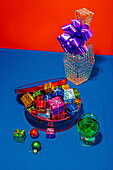 From above colorful Christmas gifts in a red tin and a sparkling vase with a purple bow on a vibrant blue table against a red background