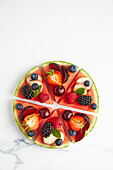 Draufsicht auf Zutaten, bestehend aus Erdbeere, Apfel, Himbeere, Trauben, Brombeere, Pflaume, Heidelbeere, Pfirsich, die auf runden, geschnittenen Scheiben auf weißer Oberfläche platziert sind, während der Zubereitung einer Wassermelonenpizza