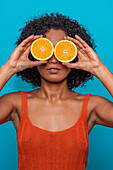 Serious young Hispanic female with curly hair covering eyes with slice of orange while standing against blue background