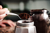 Close-up of anonymous person hand transferring freshly ground coffee from a jar to a stainless steel stovetop espresso maker set on a speckled countertop