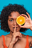 Portrait of young Hispanic female with curly black hair covering eye with orange slice and gesturing silence against blue background