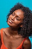 Calm positive African American female fashion model in casual wear with curly hair smiling with closed eyes against blue background