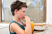 Side view of thoughtful cute boy drinking mango juice from glass and looking away seriously while sitting at table in cafe during summer vacation