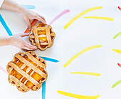 From above cropped unrecognizable hands arranging Italian Easter pastiera napoletana pastries on a white surface with colorful brushstrokes.