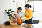 Smiling young Hispanic female friends looking down while standing at table with Halloween tools and pulling out pulp from top of orange pumpkin in daylight