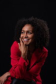 Smiling young Hispanic female model with Afro hairstyle in red coat looking away while sitting against black background