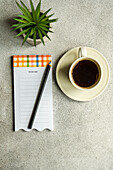 Draufsicht auf einen Kaffeebecher neben einer Topfpflanze und einem Zettel mit Bleistift auf einem grauen Tisch