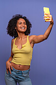 Positive Hispanic female in trendy outfit smiling and taking self portrait on mobile phone while standing against blue background