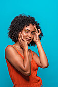 Side view of friendly African American female fashion model in casual clothes with curly hair smiling and touching face with closed eyes while standing against blue background