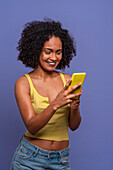 Cheerful Hispanic female with Afro hairstyle in yellow top browsing smartphone while standing on blue background