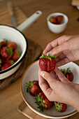 High angle of crop person hold a strawberry in hands and in bowl with green leaves over wooden dining table indoors in blurred background