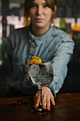 Crop female bartender looking at camera while serving wineglass with gin and tonic cocktail decorated with orange zest on wooden counter in bar