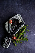 From above table setting for Christmas dinner with dark modern cutlery wrapped in green napkin with fir tree leaves and decor on concrete table