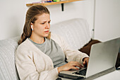 Attentive young female distance employee in earbuds typing on netbook while sitting on sofa in house room