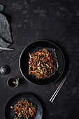 Blick von oben auf Stäbchen und Teller mit dem koreanischen Gericht Japchae, das aus Nudeln und Gemüse zubereitet wird