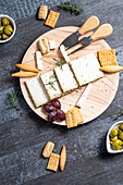 Top view of appetizing cheese served on wooden table with ripe grapes and crackers decorated by rosemary sprigs near olives in bowls on table